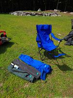 Four blue outdoor folding chairs, one set up and three packed in carry bags, placed on grass with surrounding outdoor scenery.