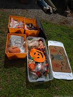 Photo showing three storage bins outdoors. One bin with orange plastic pumpkin, packaging, and decorative items. Another bin with felt Jack-o'-lantern placemat, fall wreaths, and small scarecrow figure. Third bin lid with Halloween motif placed next to bins.