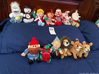 Full lot of 13 holiday plush toys laid out on a blue bedspread including characters from Rudolph the Rednosed Reindeer story