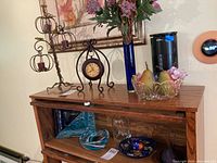 Wide view of all decor items displayed on and in wooden cabinet: art glass tray on black metal stand, blue glass vase with flowers, metal candleholder, metal clock, two different glass bowls with decorative items inside.