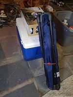 Photo showing the white and blue Gott 50-quart plastic cooler with the Wild River camp kitchen set box on top and the folded New England Patriots cooler chair in blue carry bag leaning against them.