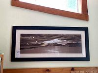 Framed black and white panoramic photo of ocean and mountainous horizon hanging on a wall beneath a window, visible reflections on glass.