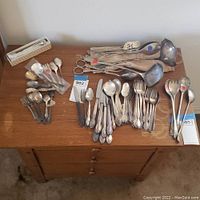 Wide view on table showing variety of silverware including spoons, forks and serving utensils in mixed condition with some tarnish and red price tags.