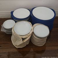 Four groups of Noritake white china on floor: large plates in blue container, smaller plates in blue container, saucers in cream bag, bowls in cream bag.
