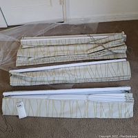 Three short curtain panels folded with visible fabric pattern and roller bars for hanging, laid on carpeted floor.