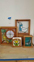 Four stained glass items on a wooden surface including a framed clock, piggy bank, cork board, and napkin holder.