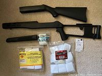 Two black synthetic rifle-style stocks, two barrel or forearm covers, two packages of cleaning patches, one small black accessory mount on beige carpet.