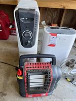 Four heating and cooling appliances arranged on a concrete floor in a garage setting. Front-to-back view of heaters and fan clustered together.