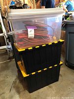 Stack of two black plastic storage bins with yellow latches, topped by a transparent plastic bin containing orange extension cords on reels inside.