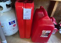 Photo showing two red plastic gas cans and one white metal propane tank. Propane tank has hazard labels and the gas cans have auction label 621 affixed.