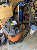 Photo of orange and black Rigid wet/dry vacuum with hose and base wheels, sitting on garage floor.