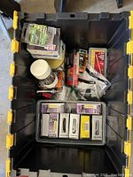 Top view inside plastic storage bin showing assorted hardware items in original packaging including multiple boxes and packs of screws and fasteners.