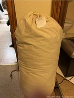 Tan fabric sack containing a rolled or packed item (sleeping bag) with drawstring closure, showing lot tag.