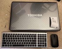 Top view of Toshiba Satellite laptop closed with visible textured cover and Toshiba logo. Wireless keyboard and mouse placed in front.