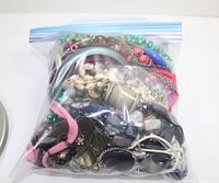 Clear plastic ziplock bag filled with various broken jewelry pieces such as chains, beads, and fabric straps, showing the volume and assortment.