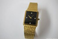 Gold tone ladies watch with woven metal bracelet and rectangular black dial. Four diamonds serve as hour markers at cardinal points on the dial.