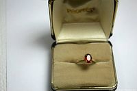 Photo of 14K yellow gold ring with oval faceted cushion set orange-red stone in cream cushioned box showing full face of the stone.