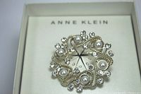 Top view of the round Anne Klein brooch showing the faux pearls and rhinestones arranged in a gold-tone metal setting, placed in its original box with Anne Klein branding visible.