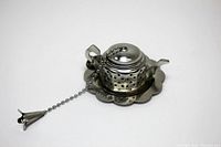 Photo of metal teapot-shaped tea infuser with handle, lid connected by ball chain, and matching draining plate