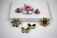 Top view of all items together including porcelain rose brooch and earrings, rhinestone flower brooches, and butterfly pins arranged on white surface.