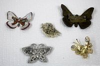 Photo showing back sides of five brooches including two butterfly shapes (one leather), one bird brooch, and two metal butterfly brooches with pin backs.