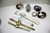 Photo of 8 different wristwatches arranged on a white surface, showing various band materials and dial styles.