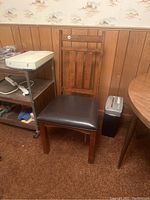 The wooden chair positioned beside a metal utility cart and near a wooden table. Shows the front and side of the chair, emphasizing the cushion and backrest design.