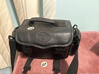Black Maycard carrying case for camcorder with handle and shoulder strap on a pink surface.