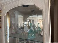 Glass display cabinet curved glass door showing clear and colored glass bottles and vases on two glass shelves.