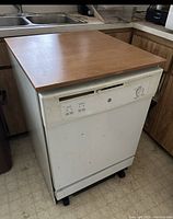 Front and left side view of the white dishwasher with wood top, showing control panel and cabinet surroundings.