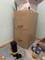Beige two-drawer metal filing cabinet placed on carpeted floor with miscellaneous items around it.