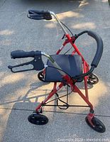 Red metal rollator walker with black cushioned seat, ergonomic handles with brakes, and four black wheels, folded and standing outdoors.