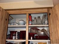 Two upper kitchen cabinets opened showing white ceramic teapot with cups, assorted glassware, red plastic cups, various plates and bowls, and plastic containers with lids.