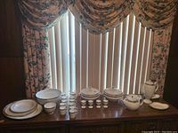 Full view of the WedgeWood Amherst white china dinnerware set arranged on a wooden surface with floral curtains in the background. Includes all plates, cups, bowls, teapot and covered serving dishes.