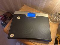 Digital bathroom scale on wooden side table, front view showing LCD display reading 0.0.