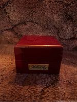 Closed maroon wooden Williamsburg box on textured brown fabric background showing metal plaque with "Williamsburg."