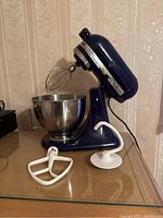 Side angled view of dark blue Kitchenaid Ultra Power mixer with stainless steel bowl and wire whisk attachment, dough hook and flat beater placed in front on table.