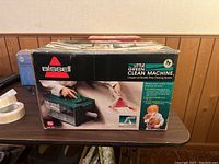 Box of Bissell Little Green Clean Machine placed on table against wood panel wall