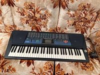 Black Casio CTK-571 electronic keyboard placed on patterned brown and orange fabric, showing full length including keys and pedal cord.