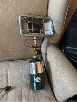 Propane lamp shown sitting on a textured fabric couch, with the lamp head protected by a metal cage and attached via valve to a green propane canister.