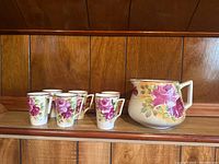 Front view of six floral ceramic cups and matching pitcher on wooden shelf.