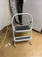 Front view of two-step white metal step ladder with black non-slip surfaces, showing curved top handle and rubber foot caps.