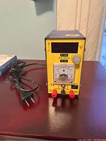 Front view of voltage meter machine showing digital voltage display, analog ammeter, voltage knob, and terminals with attached power cord.