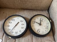 Two round wall clocks with black frames side by side on a beige fabric surface. One clock displays standard time, the other a scale to 120.