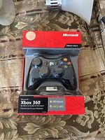 Photo showing front view of new Xbox 360 wireless controller in sealed box, highlighting Microsoft branding and compatibility with Windows and Xbox 360.