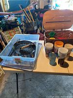 Photo showing picnic basket with plastic cups, assorted grilling tools including metal strainer pan with wooden handle and tongs placed in a clear plastic container on a table.