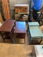 Three wooden end tables shown together: two with stained wood finish, one painted light blue, placed on a concrete floor with surrounding items.