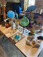 Multiple garden items arranged on white table including two gazing balls on stands, candle lanterns, shells, turtle statue, welcome sign and cherub statue.