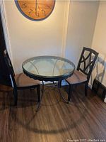 Photo showing round glass top table and two black wooden chairs with beige cushions in corner of room on wooden floor.