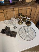 View of all items on a white table in front of wood panel background showing brass and wood sconces, candlesticks, crystal candlestick holder, rose vase, glass tray, and wood bookends.
